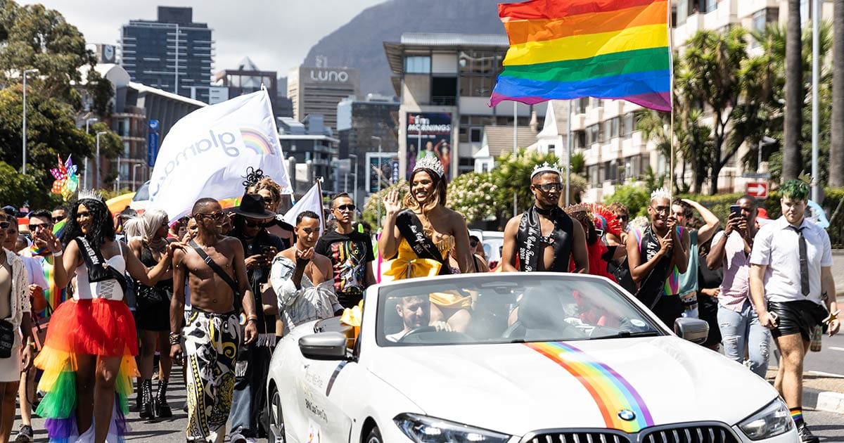 A photo of the colourful parade being led by Mr and Miss Cape Town Pride sitting in a convertible vehicle.
