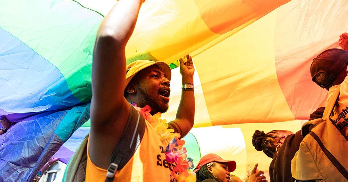A photo of a joyful man holding up a huge Pride rainbow flag.