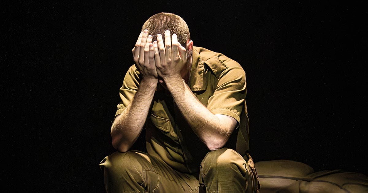 A moody photo of the stage production of Moffie, depicting a soldier sitting down and holding his face in his hands in despair.